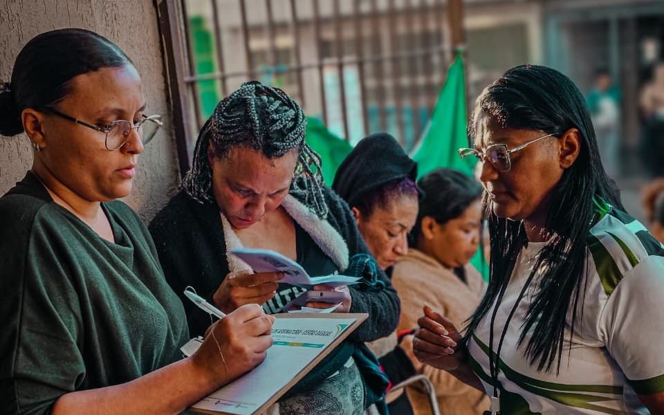 Moradoras preenchem fichas de cadastro apoiadas por uma voluntária durante um atendimento na rua.
