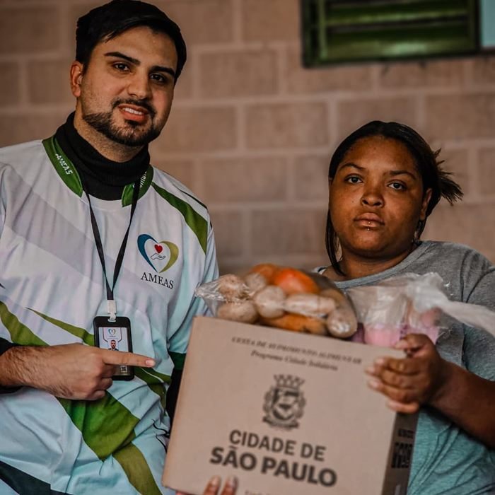 Voluntário do Instituto AMEAS e moradora seguram juntos uma cesta do programa Cidade Solidária.